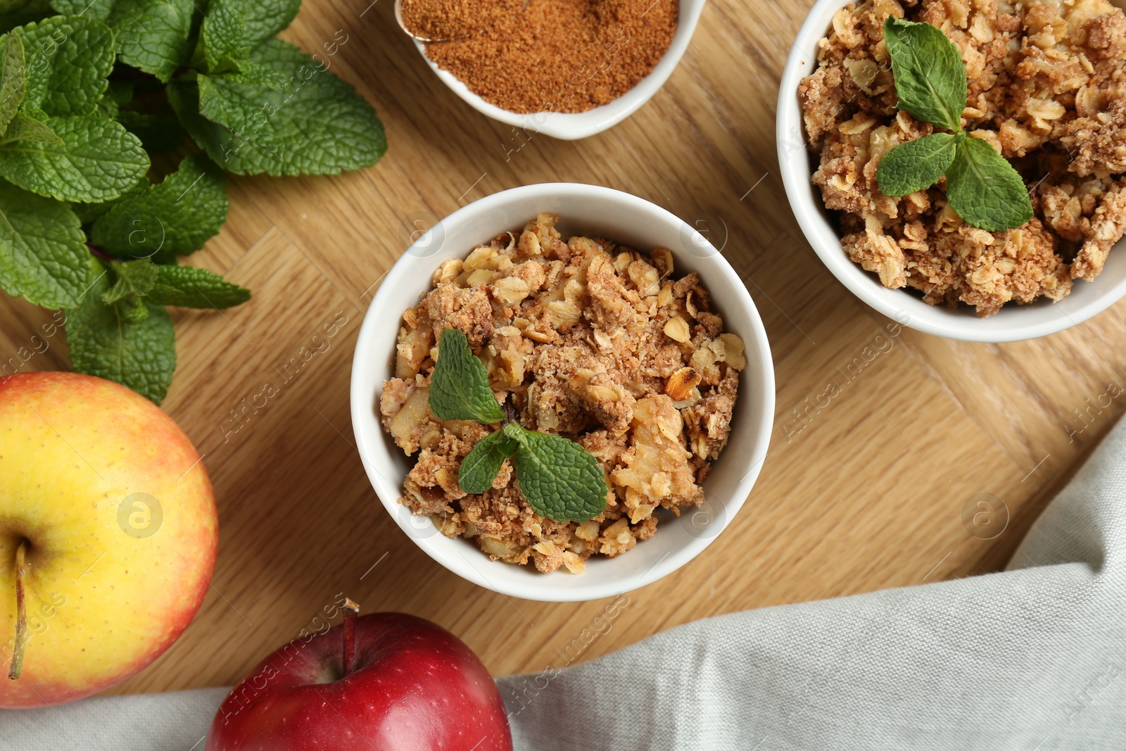 Photo of Delicious apple crisp in bowls, fresh fruits, mint and cinnamon on wooden table, flat lay