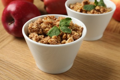 Photo of Delicious apple crisp with mint in bowls and fresh fruits on wooden table, closeup