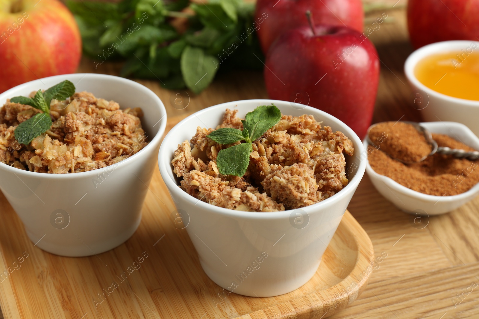 Photo of Delicious apple crisp in bowls, fresh fruits, cinnamon, mint and honey on wooden table, closeup