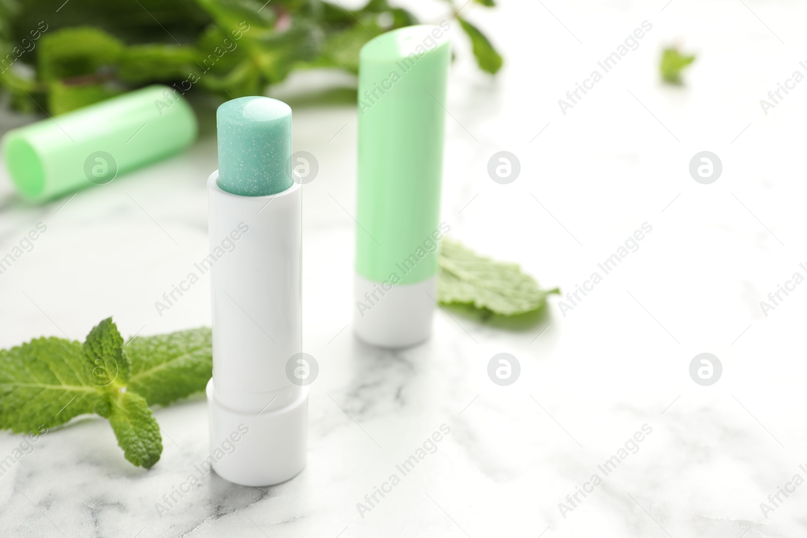 Photo of Natural lip balms and mint leaves on white marble background, closeup. Space for text