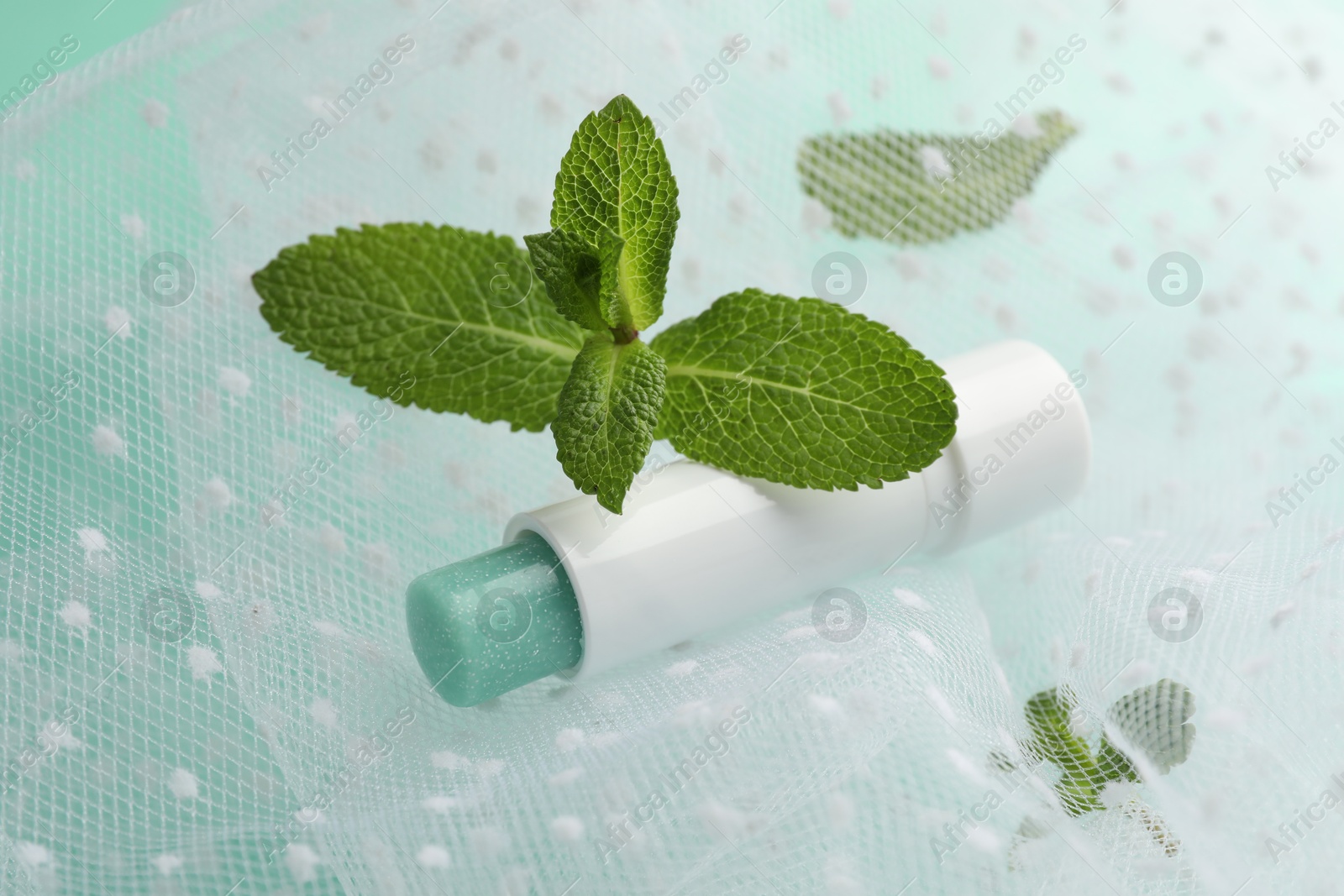 Photo of Natural lip balm, mint leaves and fabric on turquoise background, closeup