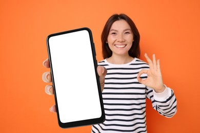 Happy woman holding mobile phone with blank screen and showing ok gesture against dark orange background