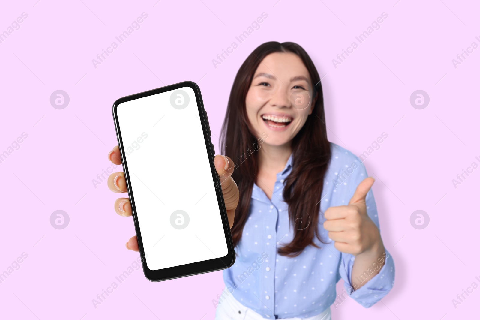 Image of Happy woman holding mobile phone with blank screen and showing thumbs-up against light violet background