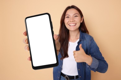 Image of Happy woman holding mobile phone with blank screen and showing thumbs-up against dark beige background