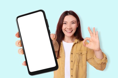 Happy woman holding mobile phone with blank screen and showing ok gesture against light blue background