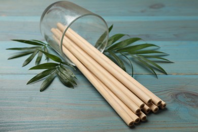 Photo of Bamboo drinking straws in glass and green leaves on light blue wooden table, closeup