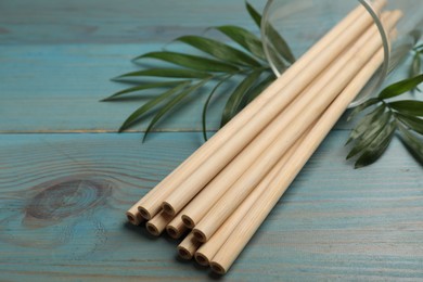 Photo of Bamboo drinking straws in glass and green leaves on light blue wooden table, closeup