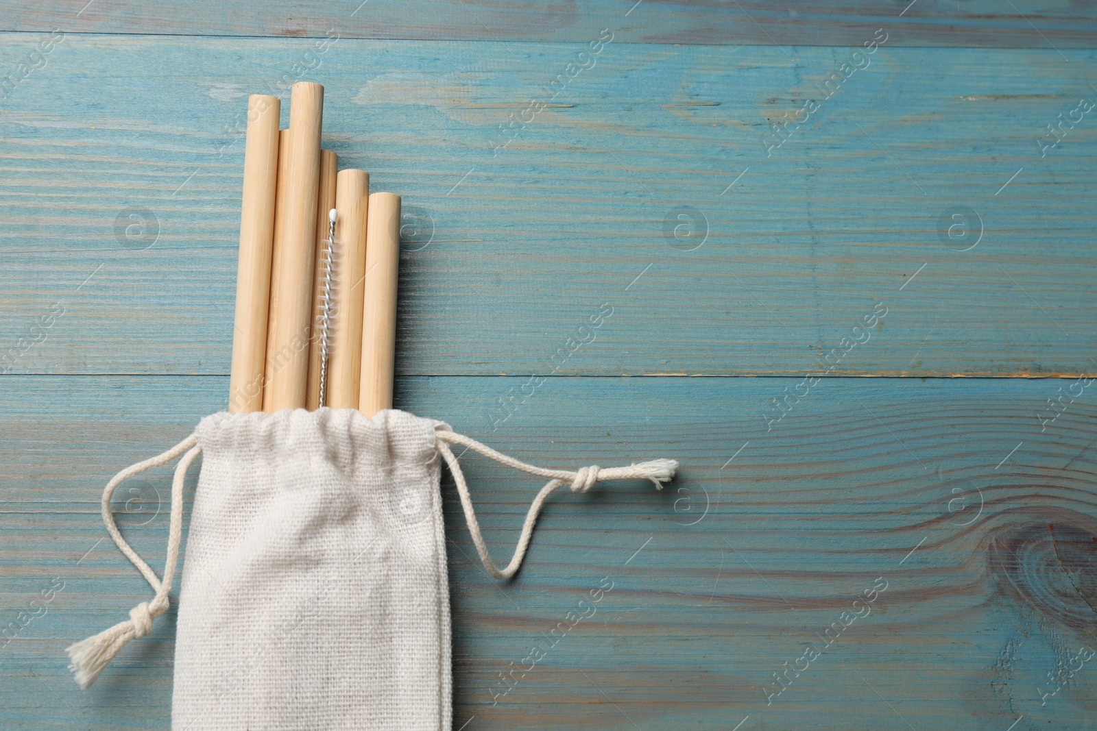 Photo of Bamboo drinking straws and cleaning brush in bag on light blue wooden table, top view. Space for text