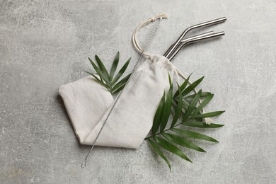 Photo of Metal drinking straws in bag, cleaning brush and green leaves on grey textured table, top view