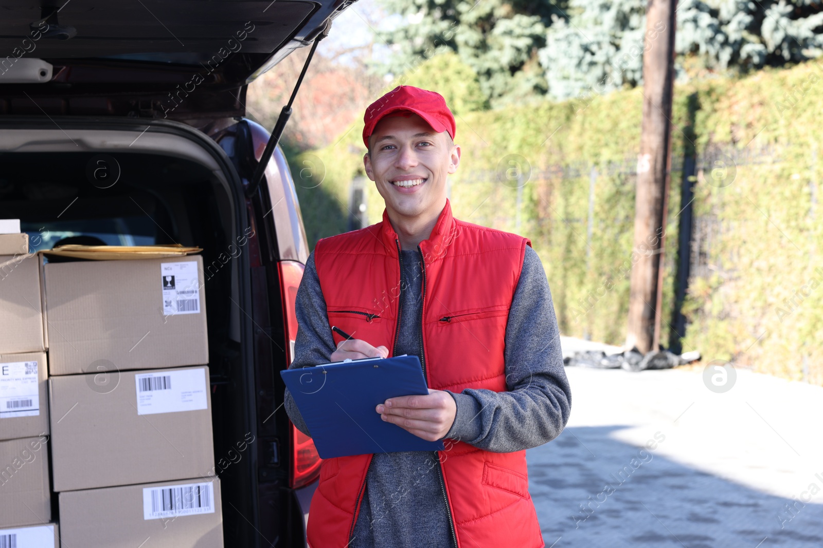 Photo of Postman with clipboard near car full of parcels outdoors