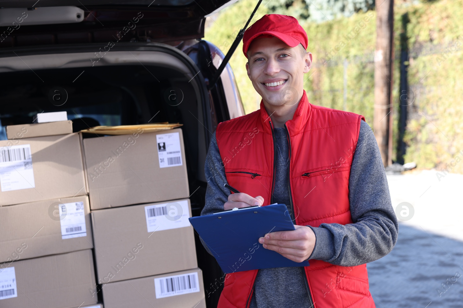 Photo of Postman with clipboard near car full of parcels outdoors