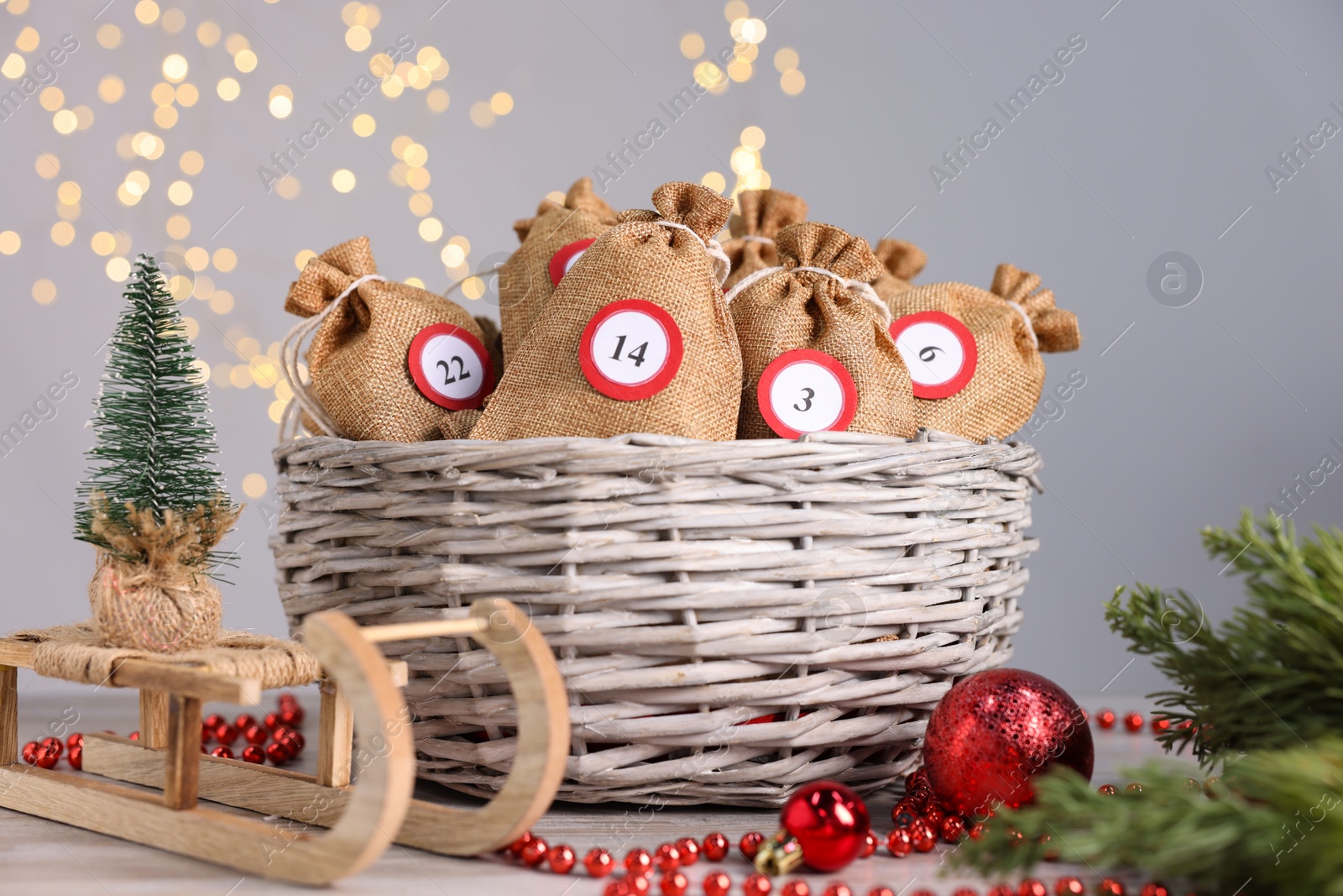 Photo of Christmas advent calendar with gifts in wicker basket and decor on table against blurred lights