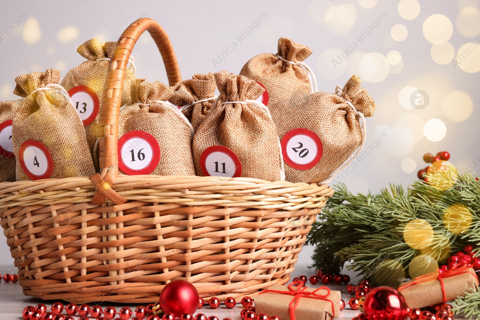 Photo of Christmas advent calendar with gifts in wicker basket and decor on table against blurred lights