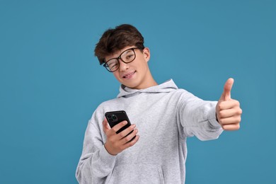 Photo of Teenage boy with smartphone showing thumbs up on light blue background