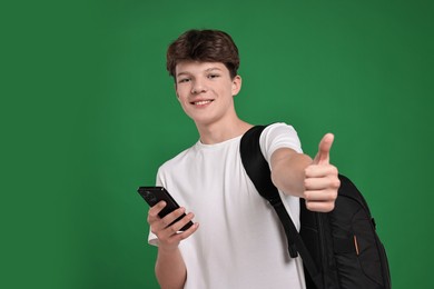 Photo of Teenage boy with smartphone and backpack showing thumbs up on green background