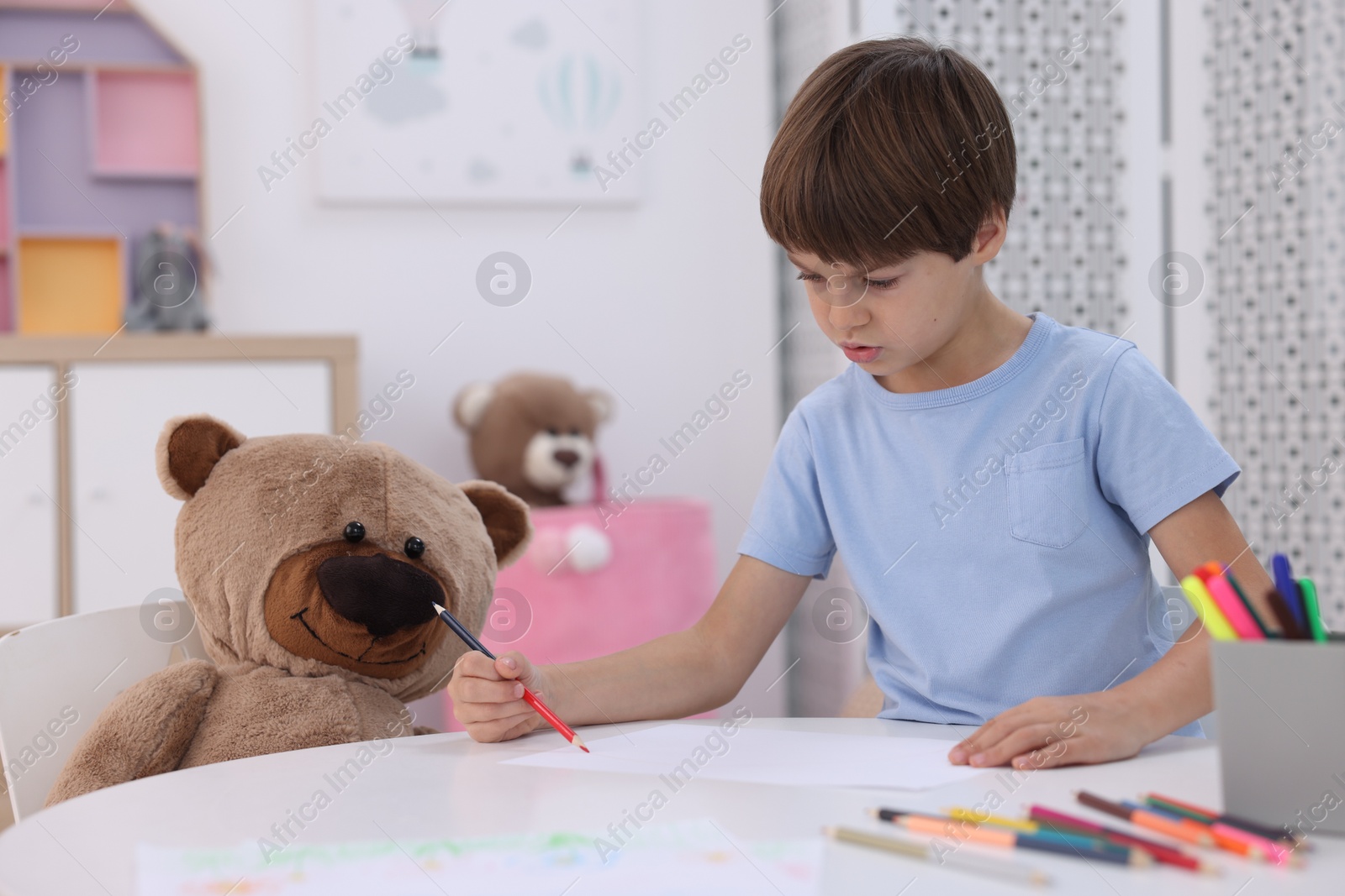 Photo of Cute boy drawing near teddy bear at white table in kindergarten