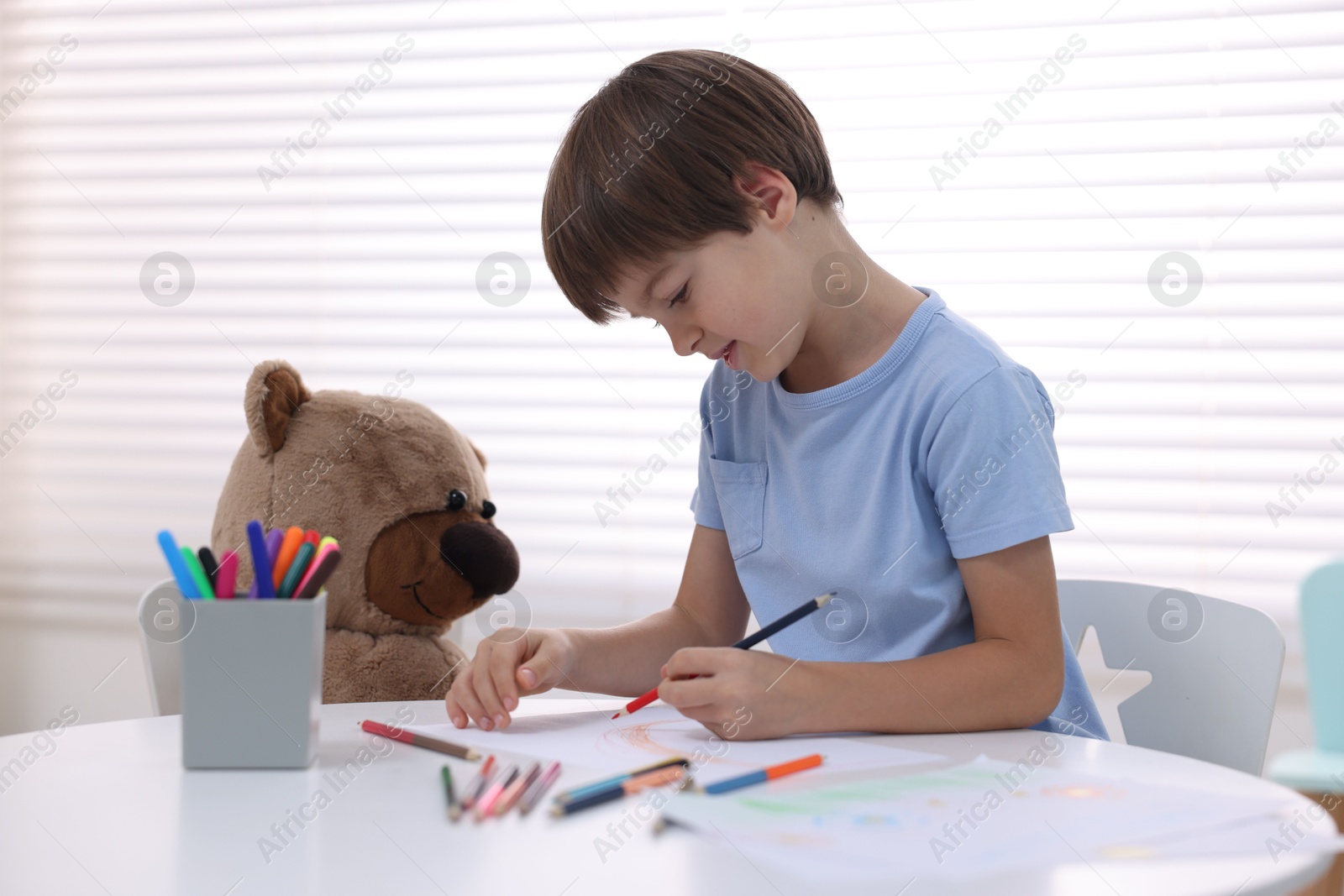 Photo of Cute boy drawing near teddy bear at white table in kindergarten