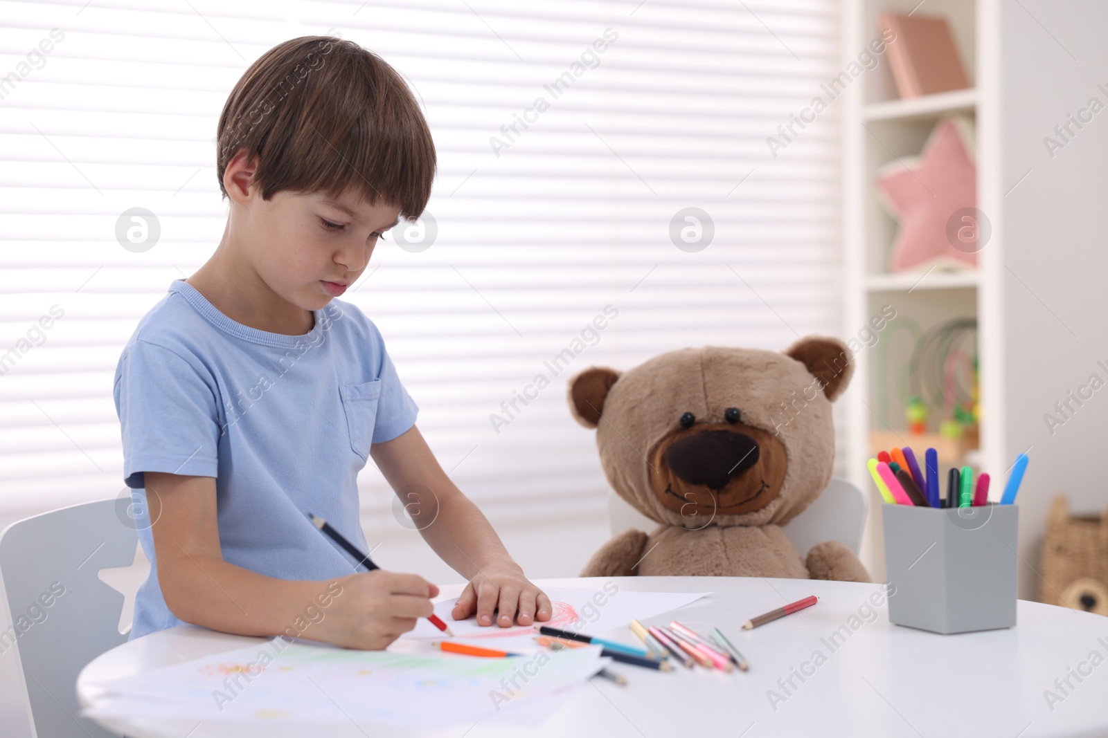 Photo of Cute boy drawing near teddy bear at white table in kindergarten