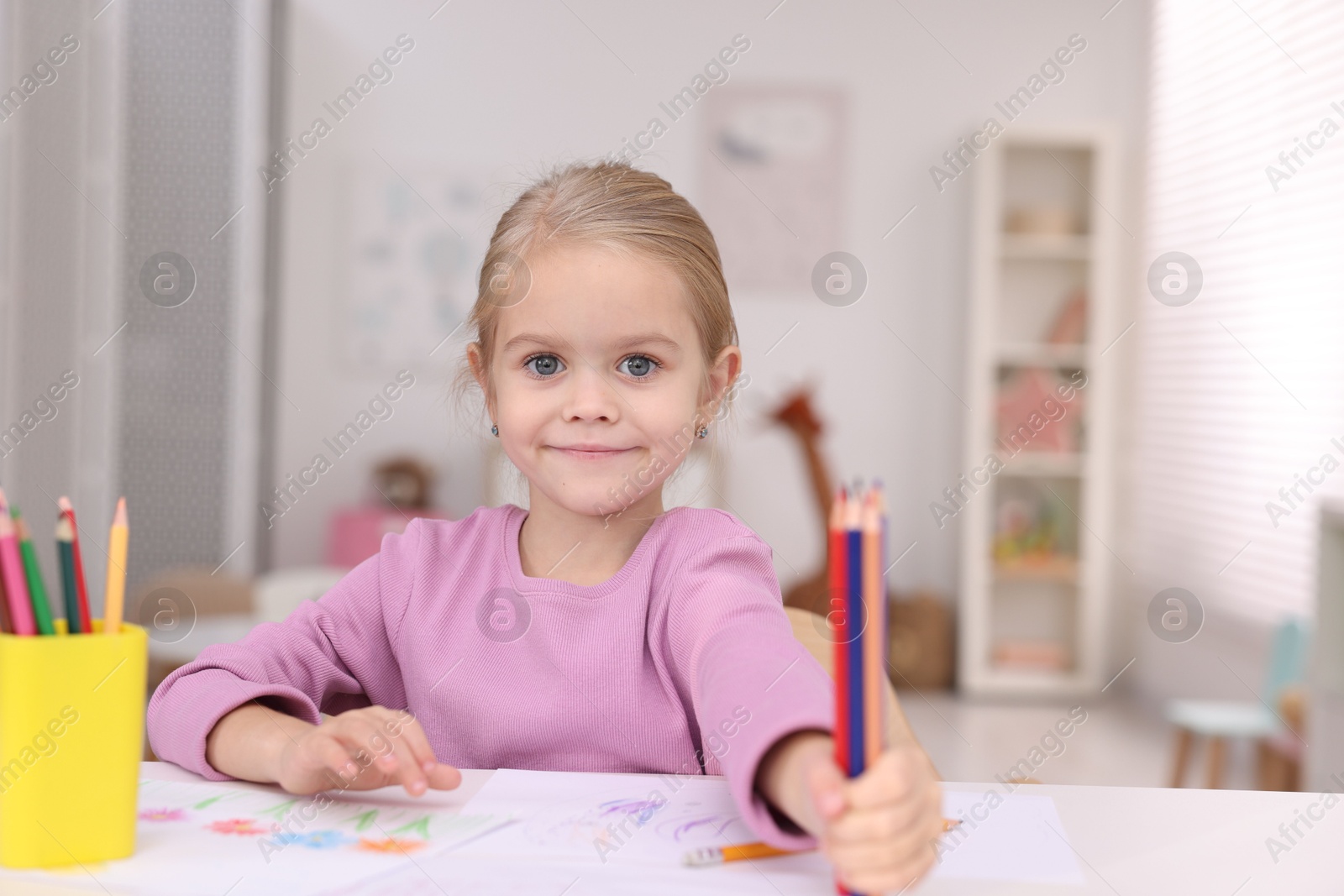 Photo of Cute girl at white table with drawing and pencils in kindergarten