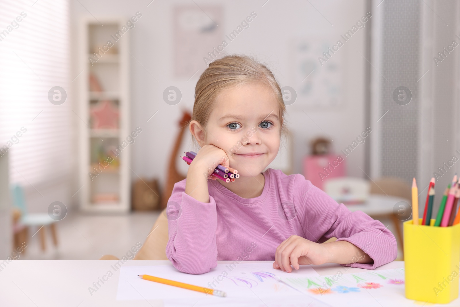 Photo of Cute girl at white table with drawing and pencils in kindergarten