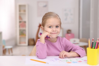 Photo of Cute girl at white table with drawing and pencils in kindergarten