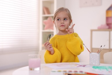 Photo of Cute girl thinking about her drawing at white table in kindergarten. Space for text