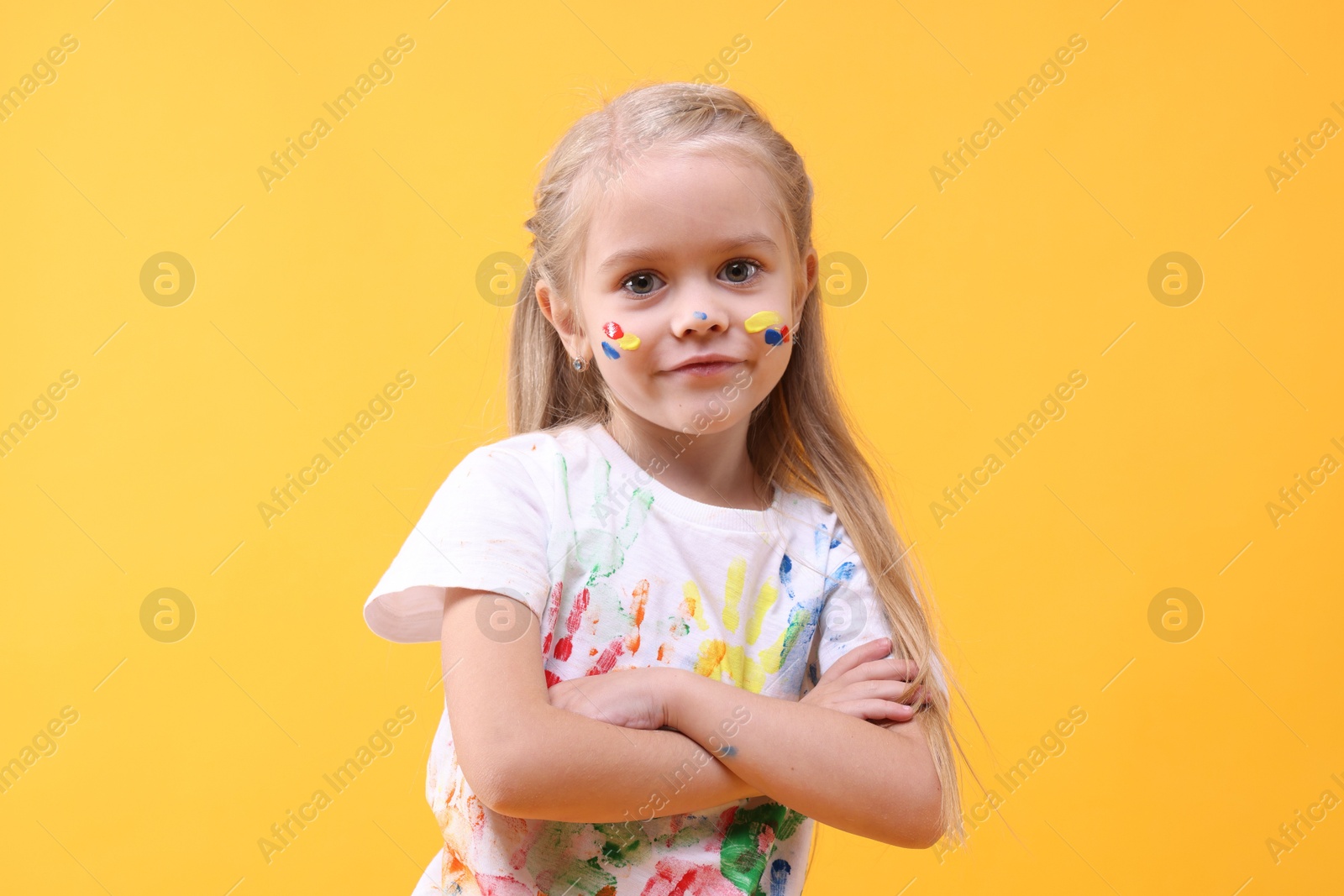 Photo of Cute girl with crossed arms smeared in paint on orange background