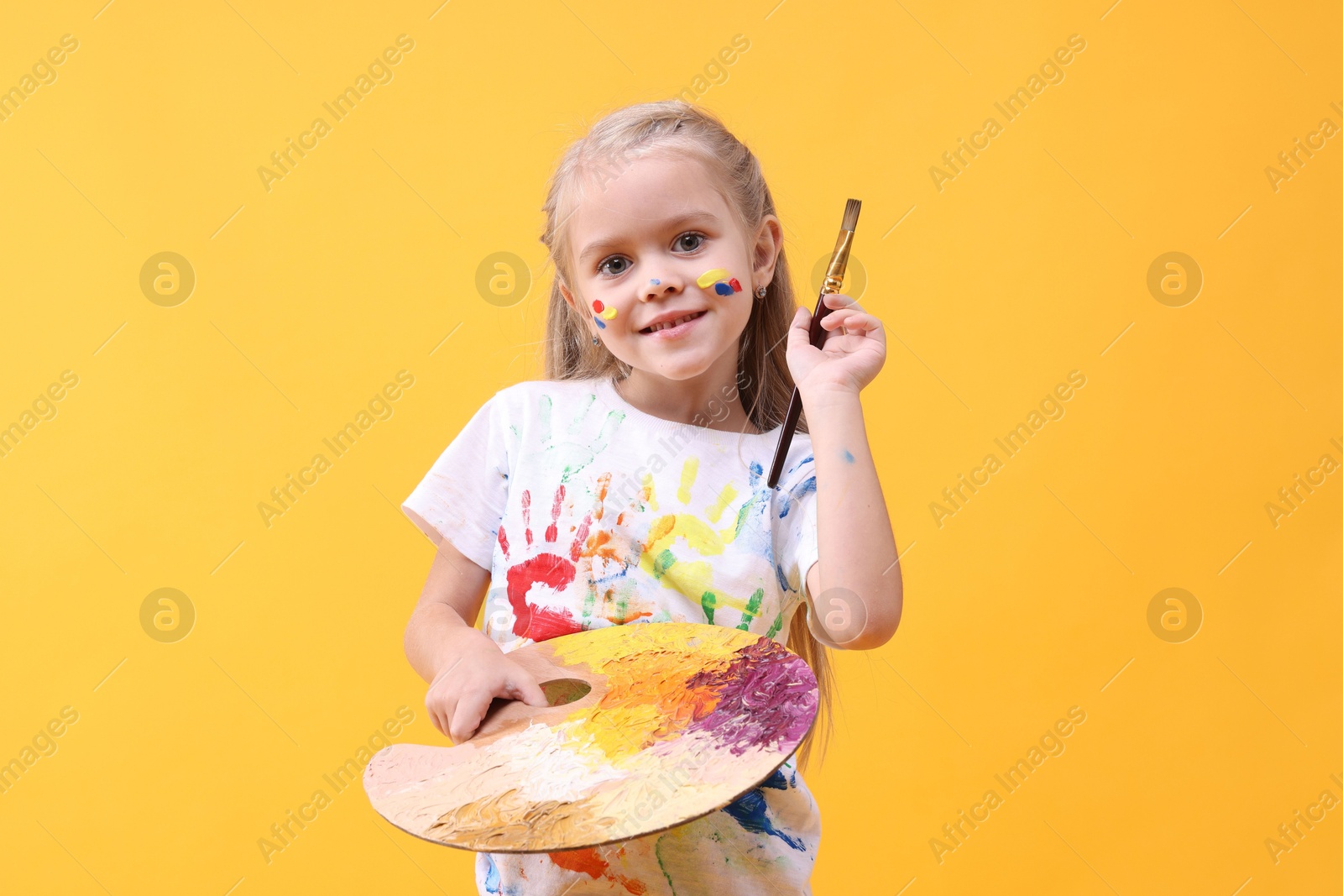 Photo of Smiling girl with brush and palette smeared in paint on orange background