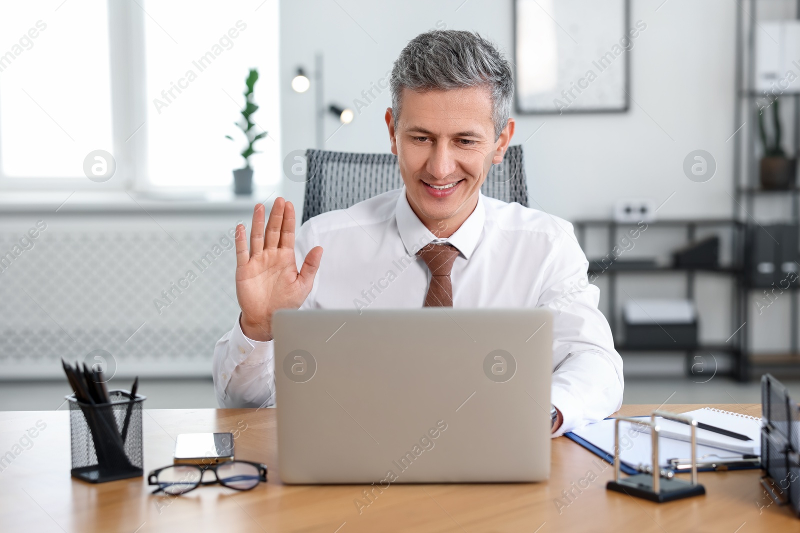 Photo of Smiling middle aged man having videochat via laptop at table in office