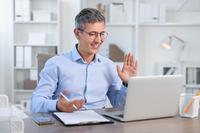 Photo of Smiling middle aged man having videochat via laptop at table in office