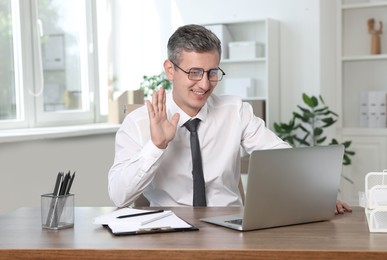 Photo of Smiling middle aged man having videochat via laptop at table in office