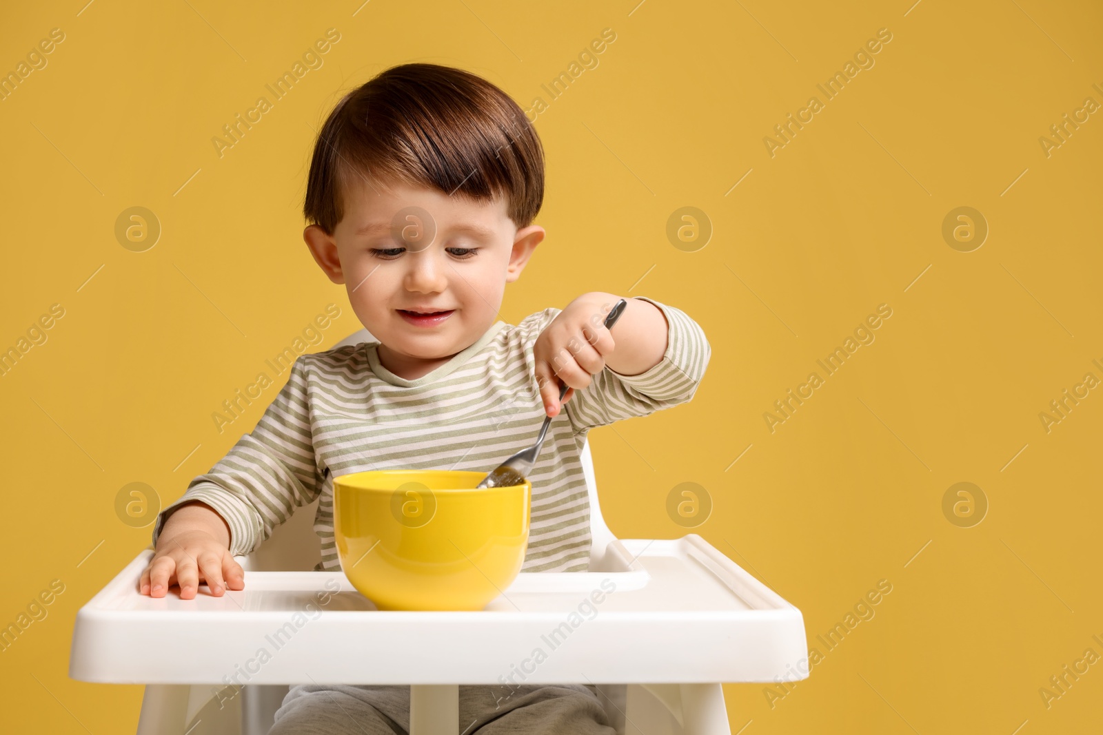 Photo of Cute little kid eating healthy baby food from bowl in high chair on yellow background