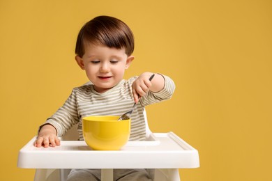 Cute little kid eating healthy baby food from bowl in high chair on yellow background