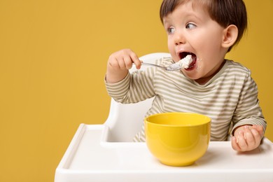 Photo of Cute little kid eating healthy baby food from bowl in high chair on yellow background, space for text