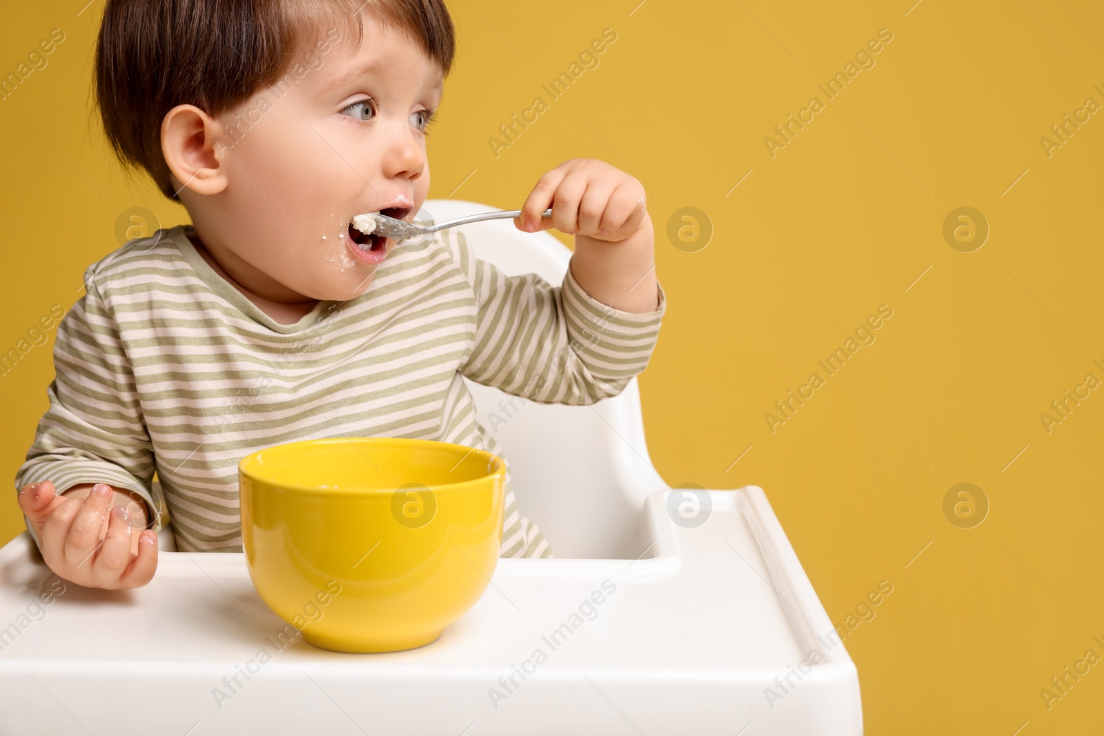 Photo of Cute little kid eating healthy baby food from bowl in high chair on yellow background