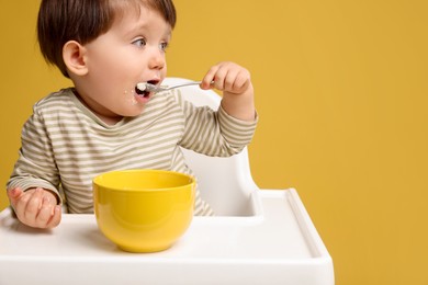 Photo of Cute little kid eating healthy baby food from bowl in high chair on yellow background