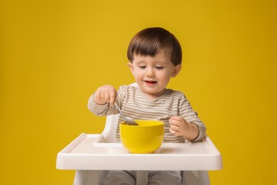 Photo of Cute little kid eating healthy baby food from bowl in high chair on yellow background