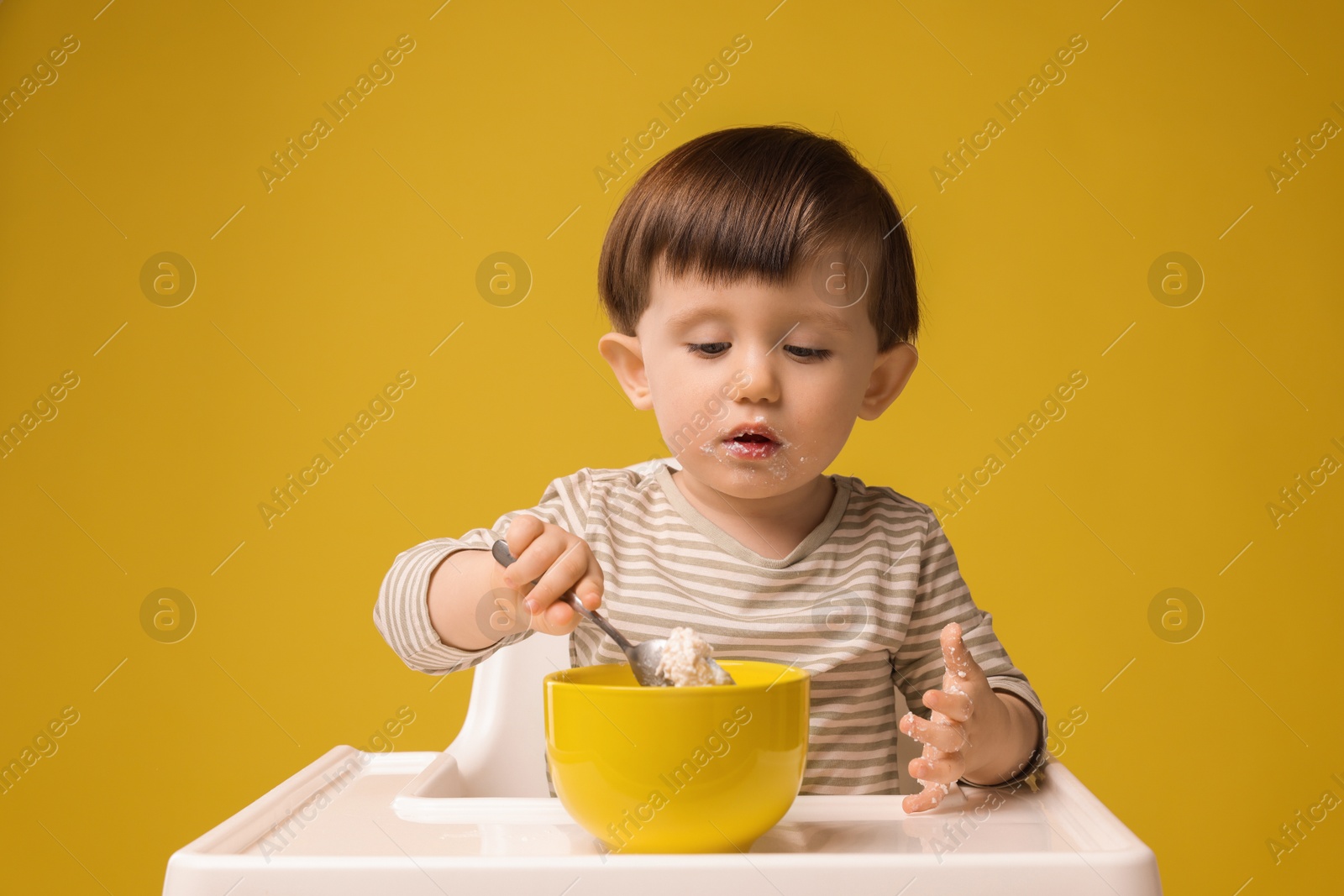 Photo of Cute little kid eating healthy baby food from bowl in high chair on yellow background