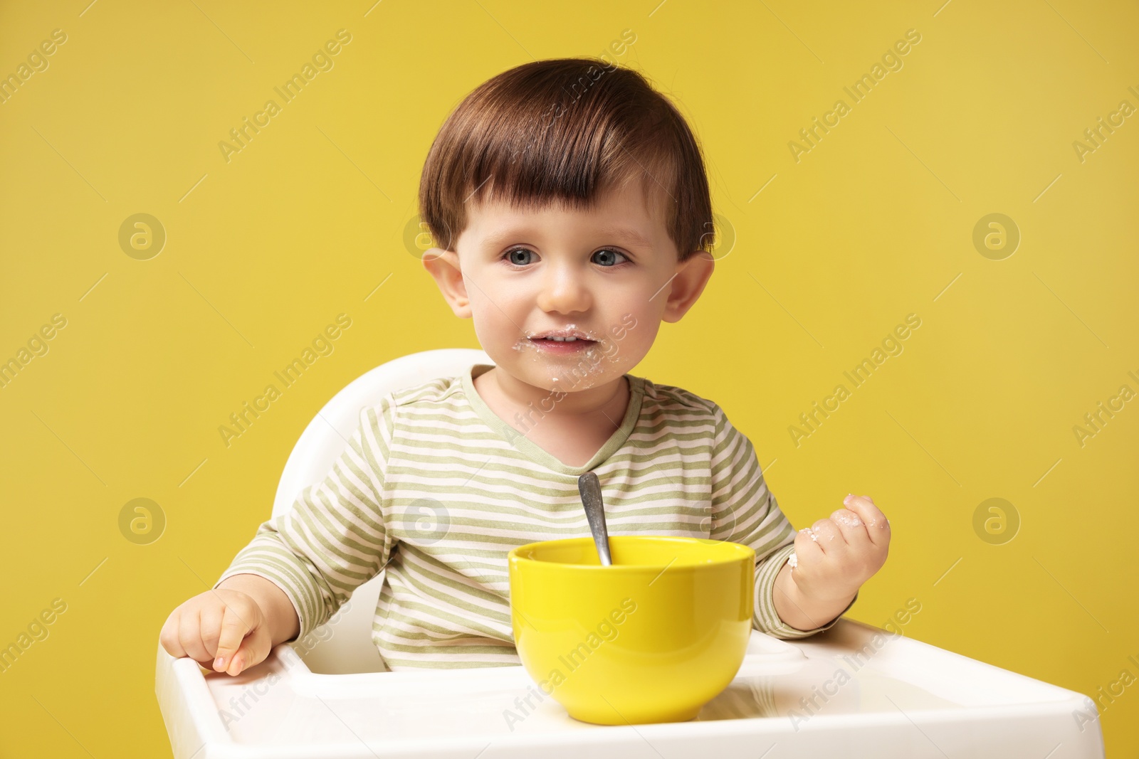 Photo of Cute little kid eating healthy baby food from bowl in high chair on yellow background