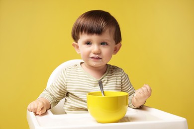 Photo of Cute little kid eating healthy baby food from bowl in high chair on yellow background