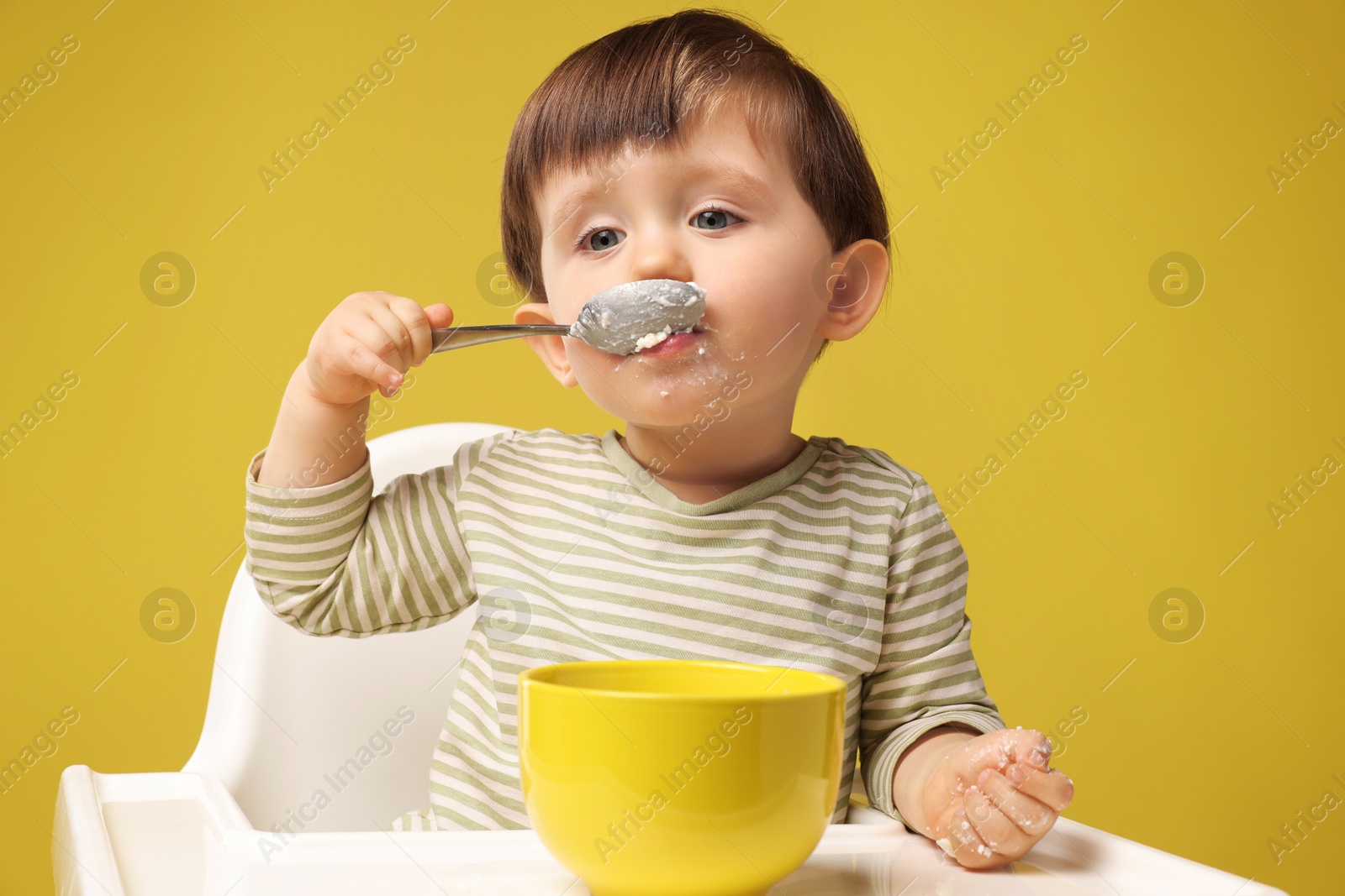 Photo of Cute little kid eating healthy baby food from bowl in high chair on yellow background