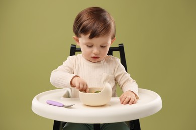 Photo of Cute little baby eating healthy food from bowl in high chair on olive background