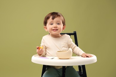 Photo of Cute little baby eating healthy food from bowl in high chair on olive background