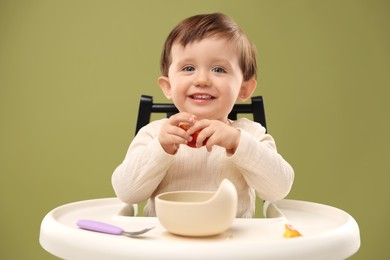 Cute little baby eating healthy food from bowl in high chair on olive background