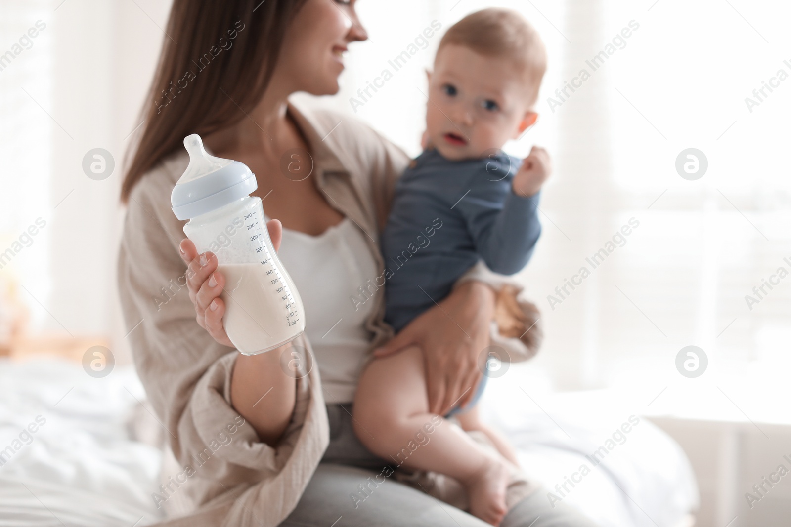 Photo of Mother holding cute little baby and bottle of milk on bed at home, selective focus