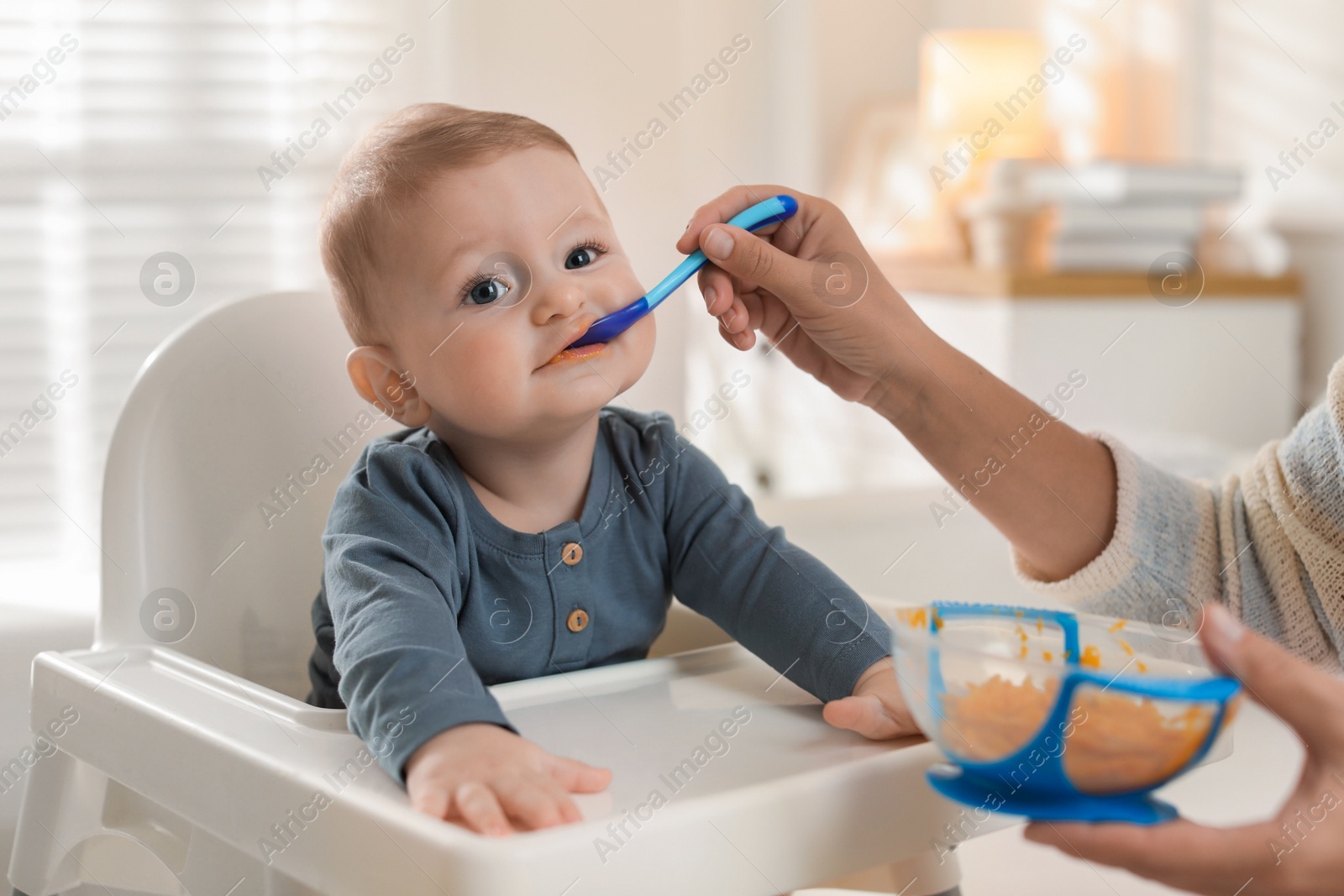 Photo of Mother feeding her cute little baby in high chair at home, closeup