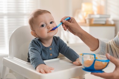 Photo of Mother feeding her cute little baby in high chair at home, closeup