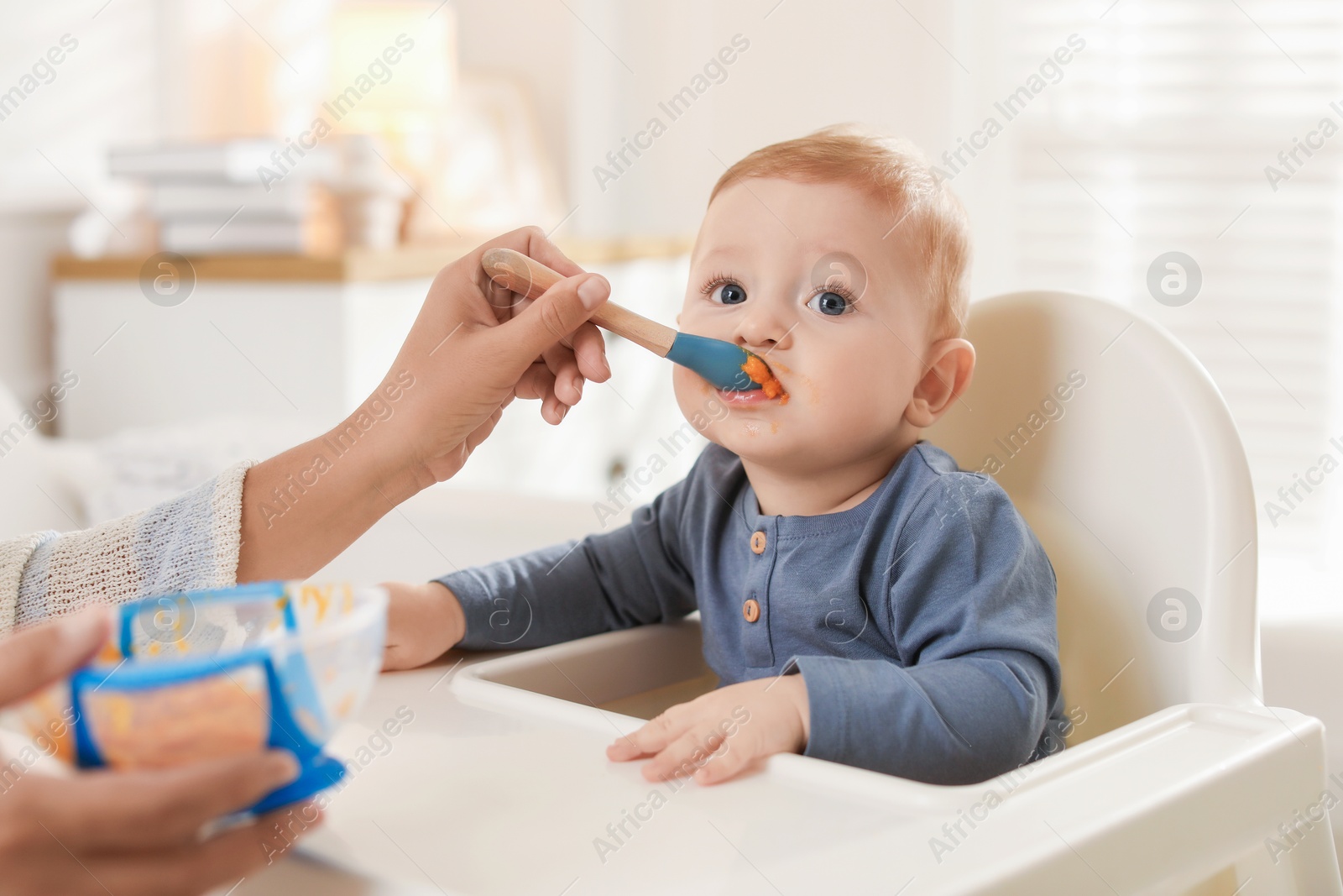 Photo of Mother feeding her cute little baby in high chair at home, closeup