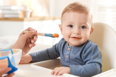 Photo of Mother feeding her cute little baby in high chair at home, closeup