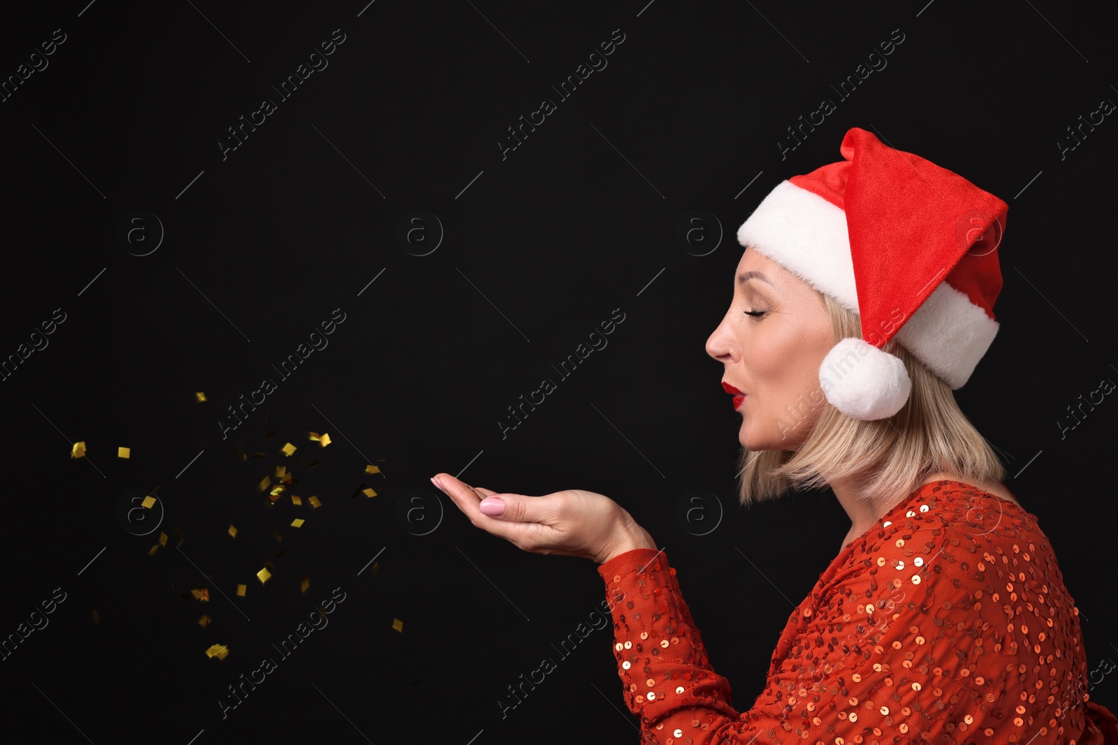 Photo of Woman with perfect makeup in Santa hat blowing confetti on black background. Christmas celebration